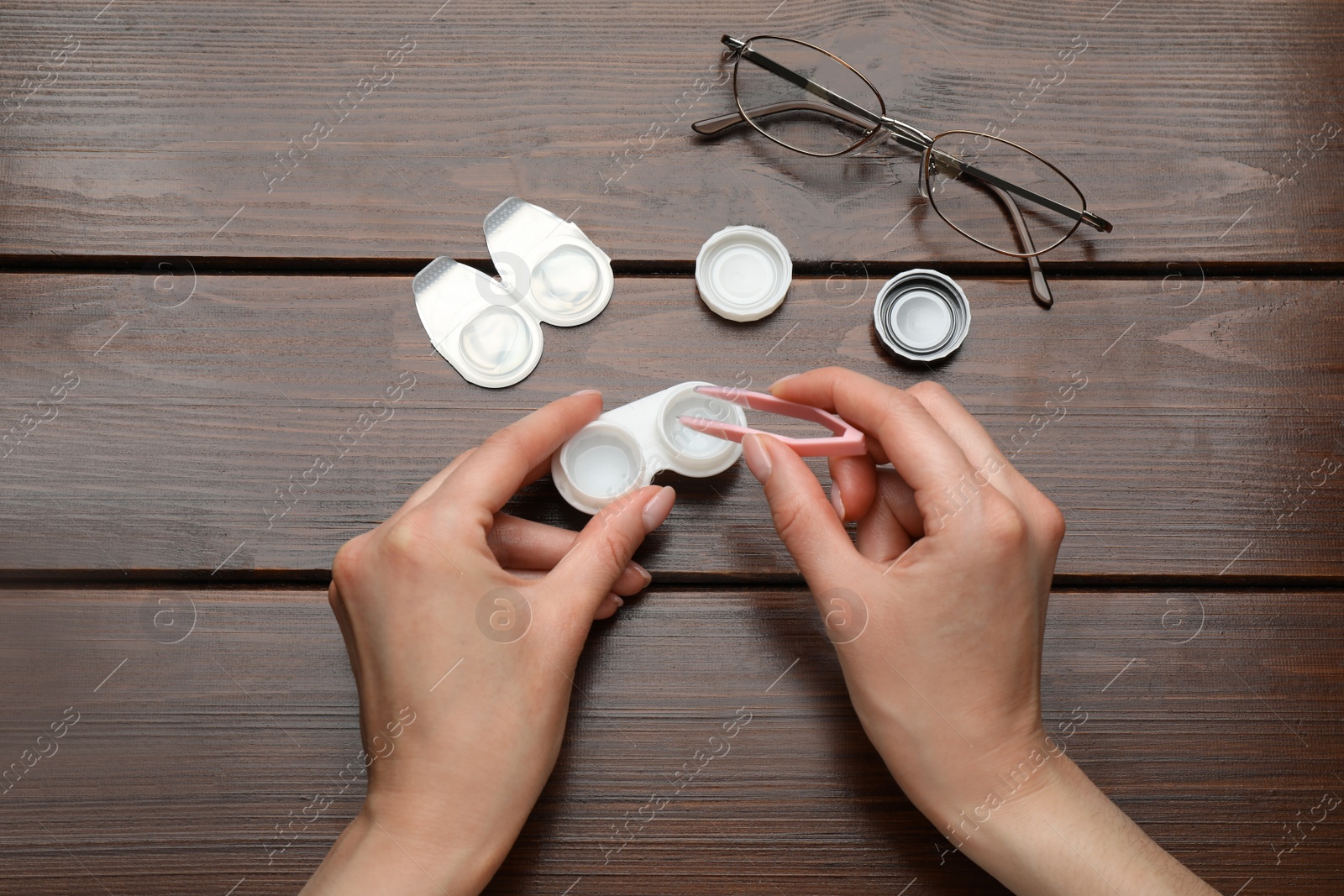 Photo of Woman taking contact lens from case with tweezers at wooden table, top view