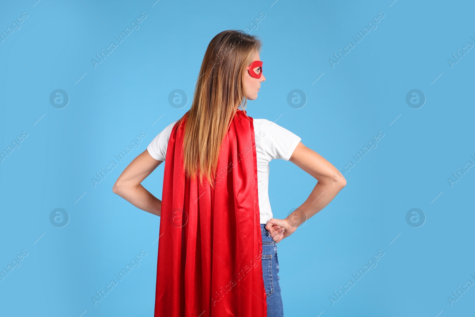 Photo of Woman wearing superhero cape and mask on light blue background, back view