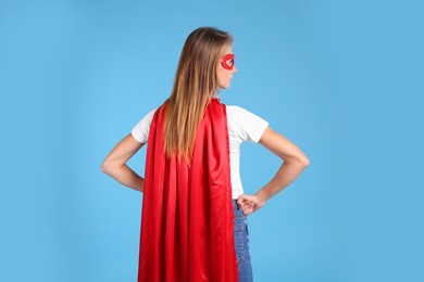 Woman wearing superhero cape and mask on light blue background, back view