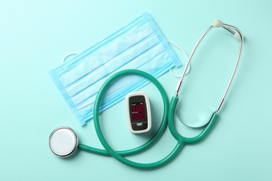 Flat lay composition with modern fingertip pulse oximeter, mask and stethoscope on light blue background