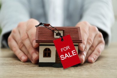 Real estate agent holding house model with SALE label at wooden table, closeup