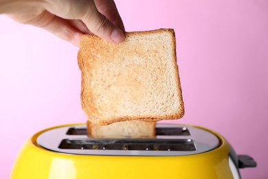 Woman taking off roasted bread from toaster against pale pink background, closeup
