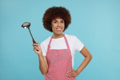 Photo of Happy young woman in apron holding ladle on light blue background