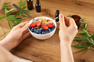 Woman dripping THC tincture or CBD oil into oatmeal bowl at wooden table, closeup