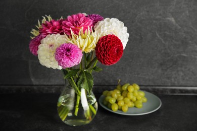 Photo of Bouquet of beautiful Dahlia flowers in vase and plate with grapes on black table near grey wall