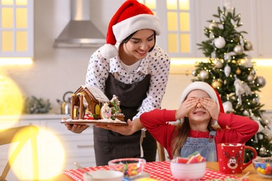 Mother surprising her daughter with gingerbread house indoors