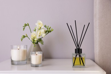Photo of Aromatic reed air freshener, freesia flowers and candles on white bedside table indoors