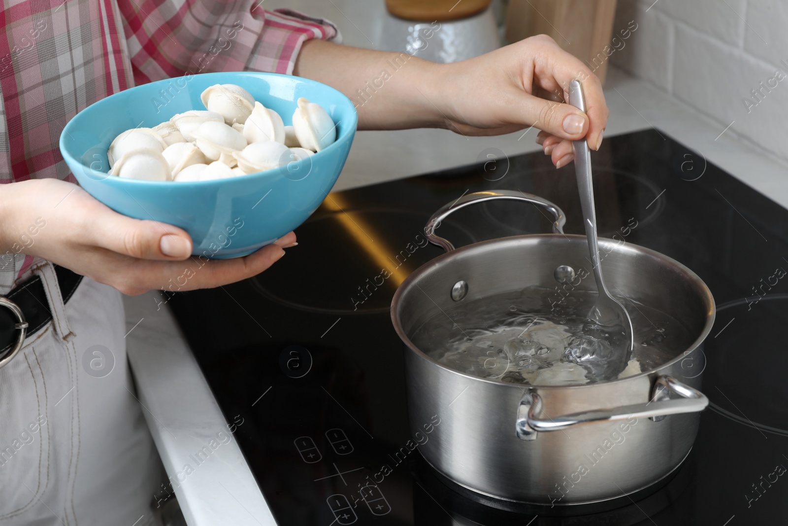 Photo of Woman putting frozen dumplings into saucepan with boiling water on cooktop in kitchen, closeup