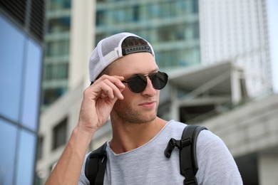 Photo of Handsome young man with stylish sunglasses and backpack near building outdoors, space for text