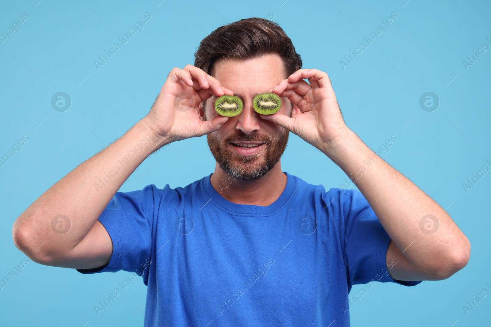 Photo of Man holding halves of kiwi near his eyes on light blue background