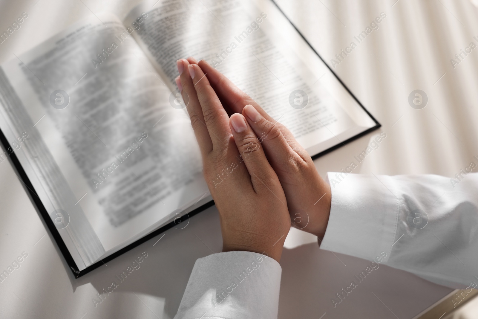 Photo of Woman holding hands clasped while praying over Bible at white table, closeup