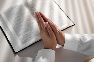 Photo of Woman holding hands clasped while praying over Bible at white table, closeup