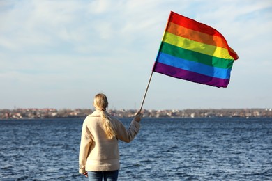 Photo of Woman holding bright LGBT flag near river, back view