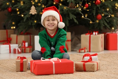 Photo of Little child wearing Santa hat with gift boxes on floor near Christmas tree at home