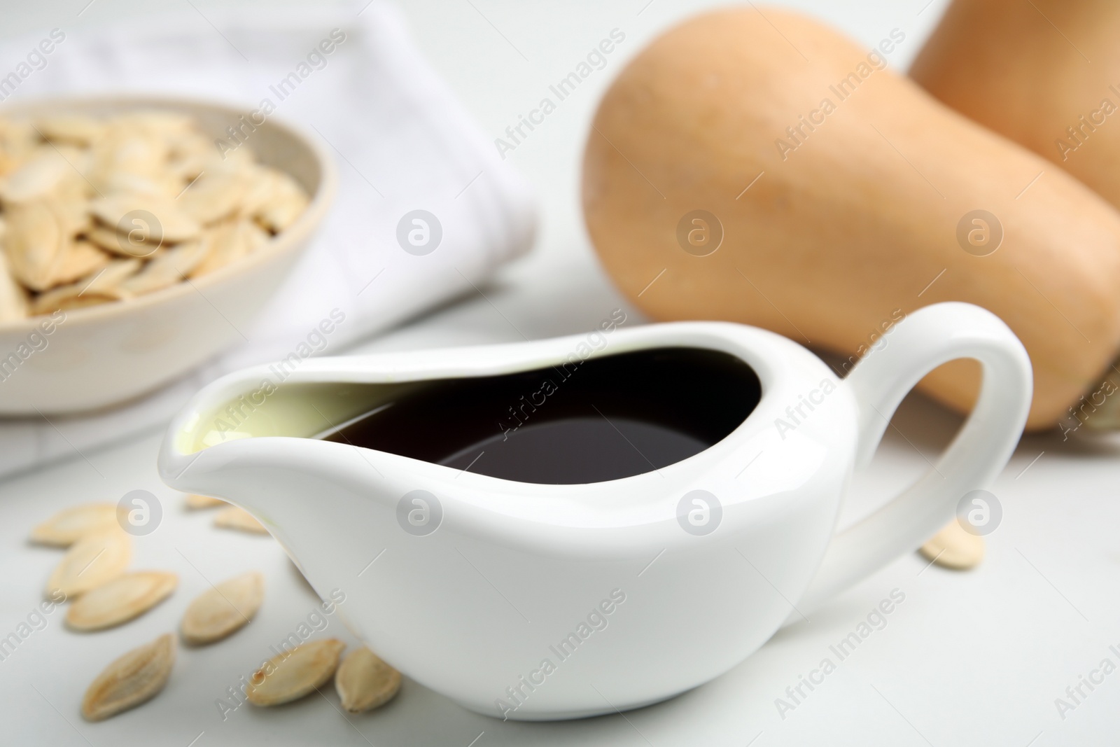 Photo of Gravy boat of pumpkin oil and seeds on white table, closeup