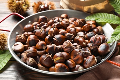 Photo of Delicious roasted edible chestnuts in frying pan on wooden table, closeup