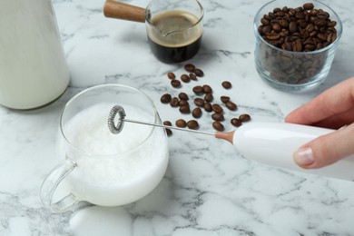 Woman whisking milk in cup with mini mixer (milk frother) at white marble table, closeup