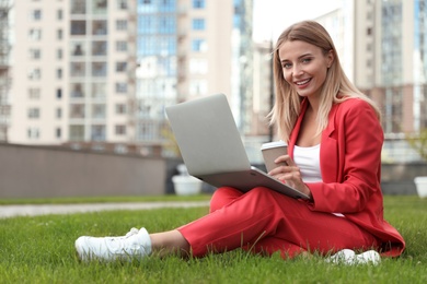 Beautiful businesswoman with laptop and coffee sitting on green lawn outdoors