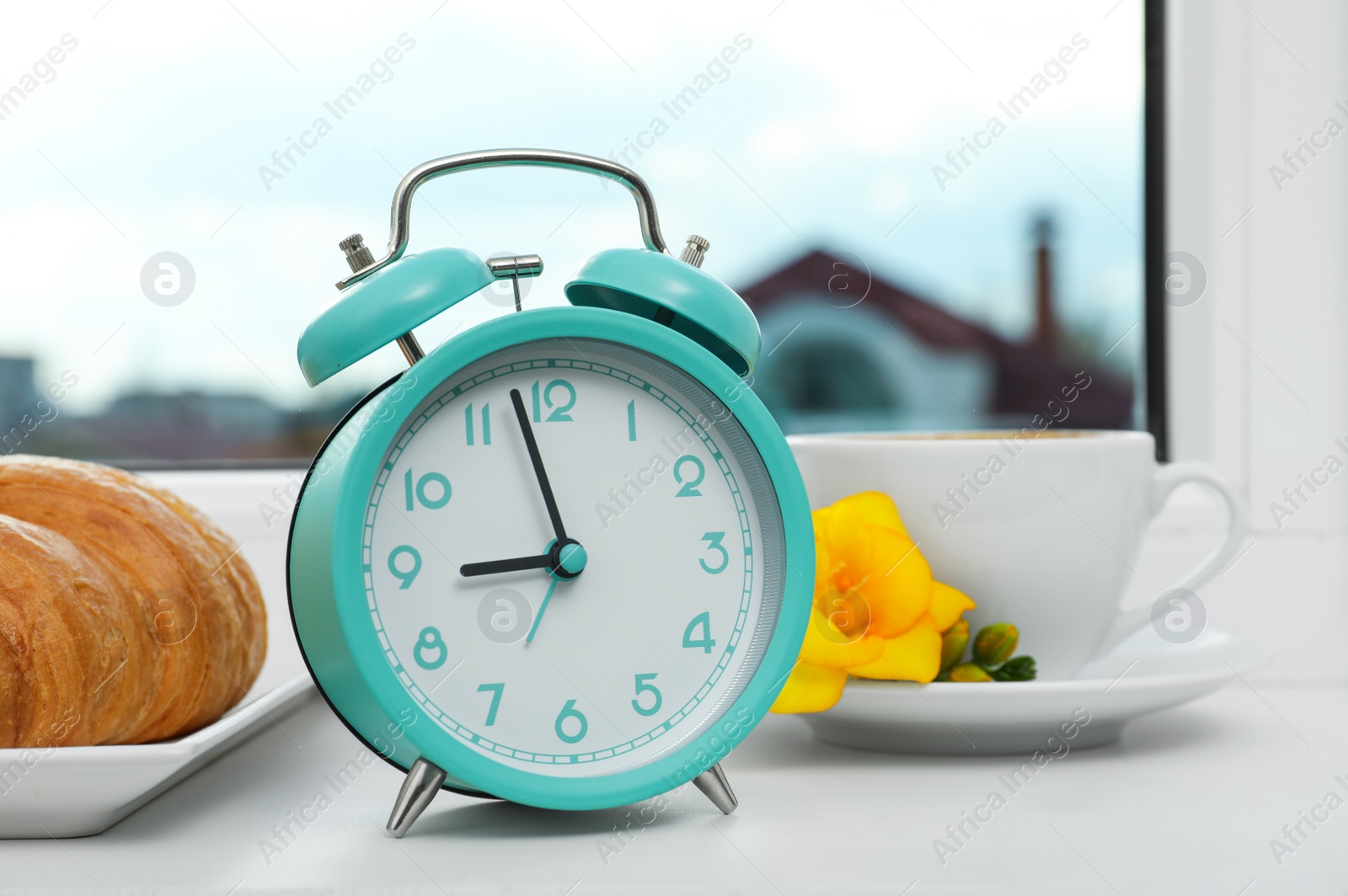 Photo of Alarm clock, croissant and cup of drink with flower on windowsill. Good morning