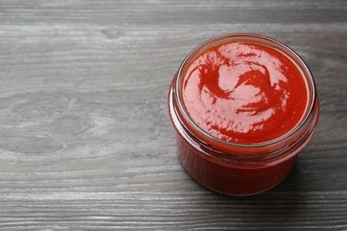 Photo of Tasty ketchup in jar on grey wooden table, space for text. Tomato sauce