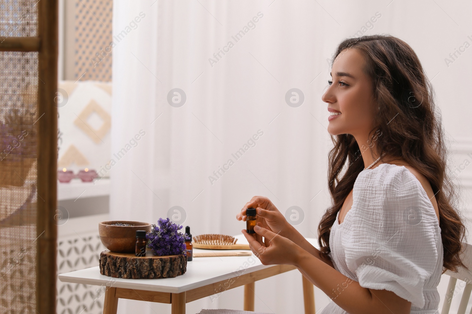 Photo of Beautiful young woman with bottle of essential oil at table in room