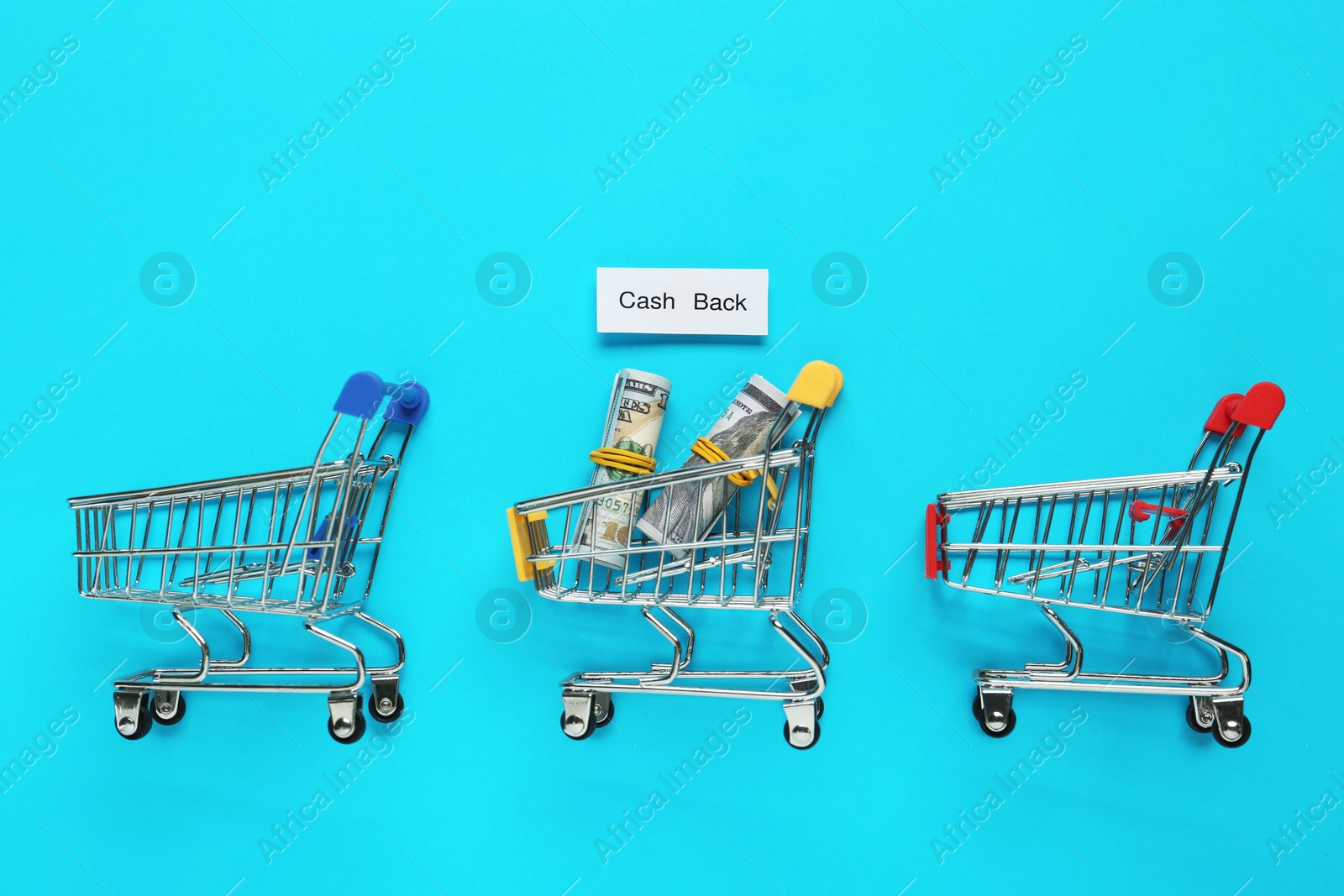 Photo of Card with word Cashback, rolled dollar banknotes and shopping carts on light blue background, flat lay