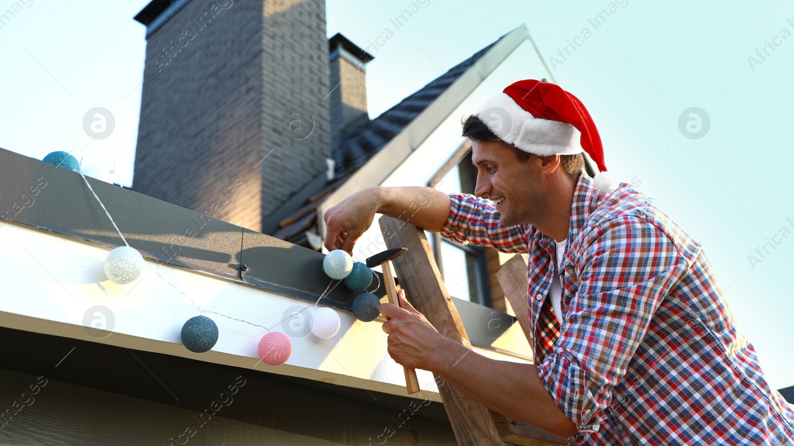 Photo of Man in Santa hat decorating house with Christmas lights outdoors