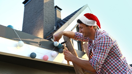 Man in Santa hat decorating house with Christmas lights outdoors