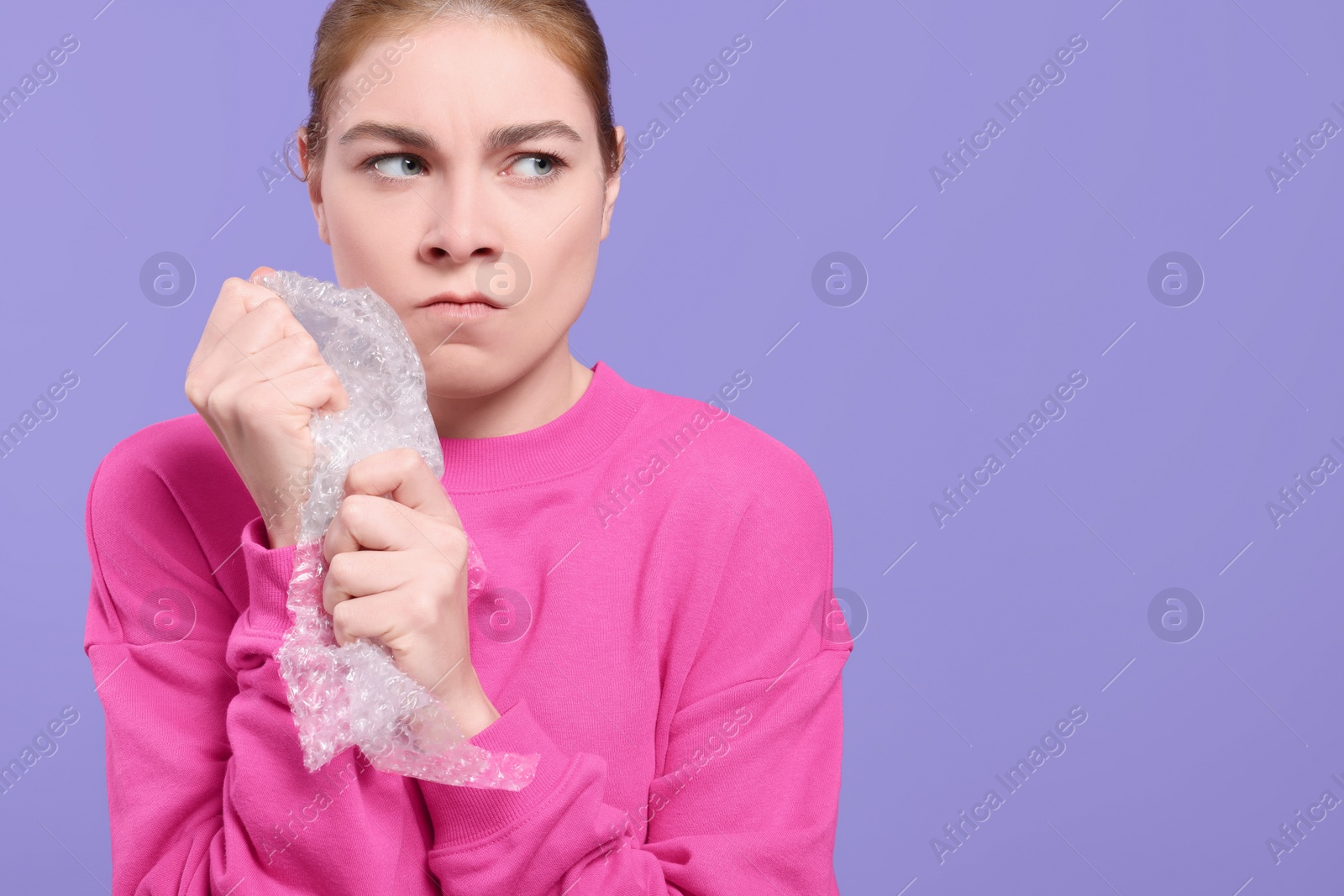 Photo of Angry woman popping bubble wrap on purple background, space for text. Stress relief