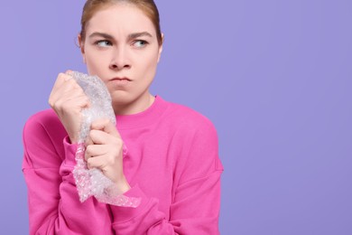 Angry woman popping bubble wrap on purple background, space for text. Stress relief