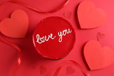 Photo of Bento cake with text Love You, ribbon and paper hearts on red table, flat lay. St. Valentine's day surprise