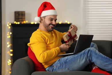 Celebrating Christmas online with exchanged by mail presents. Happy man in Santa hat with gift box during video call on laptop at home