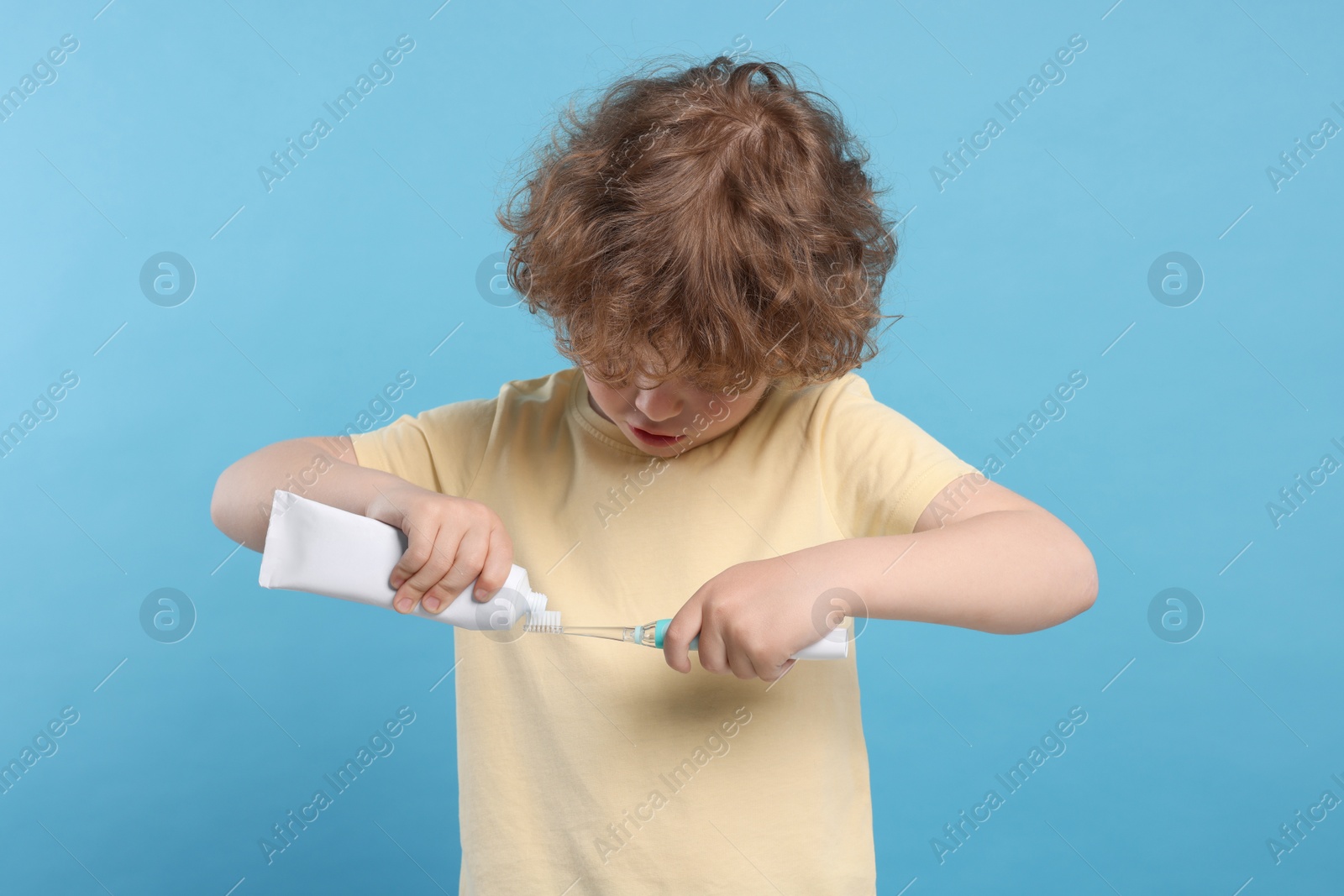 Photo of Cute little boy squeezing toothpaste from tube onto electric toothbrush on light blue background