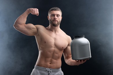 Young man with muscular body holding jar of protein powder on dark grey background