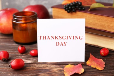 Photo of Paper card with words Thanksgiving Day near autumn leaves, berries, books and candle on wooden table, closeup