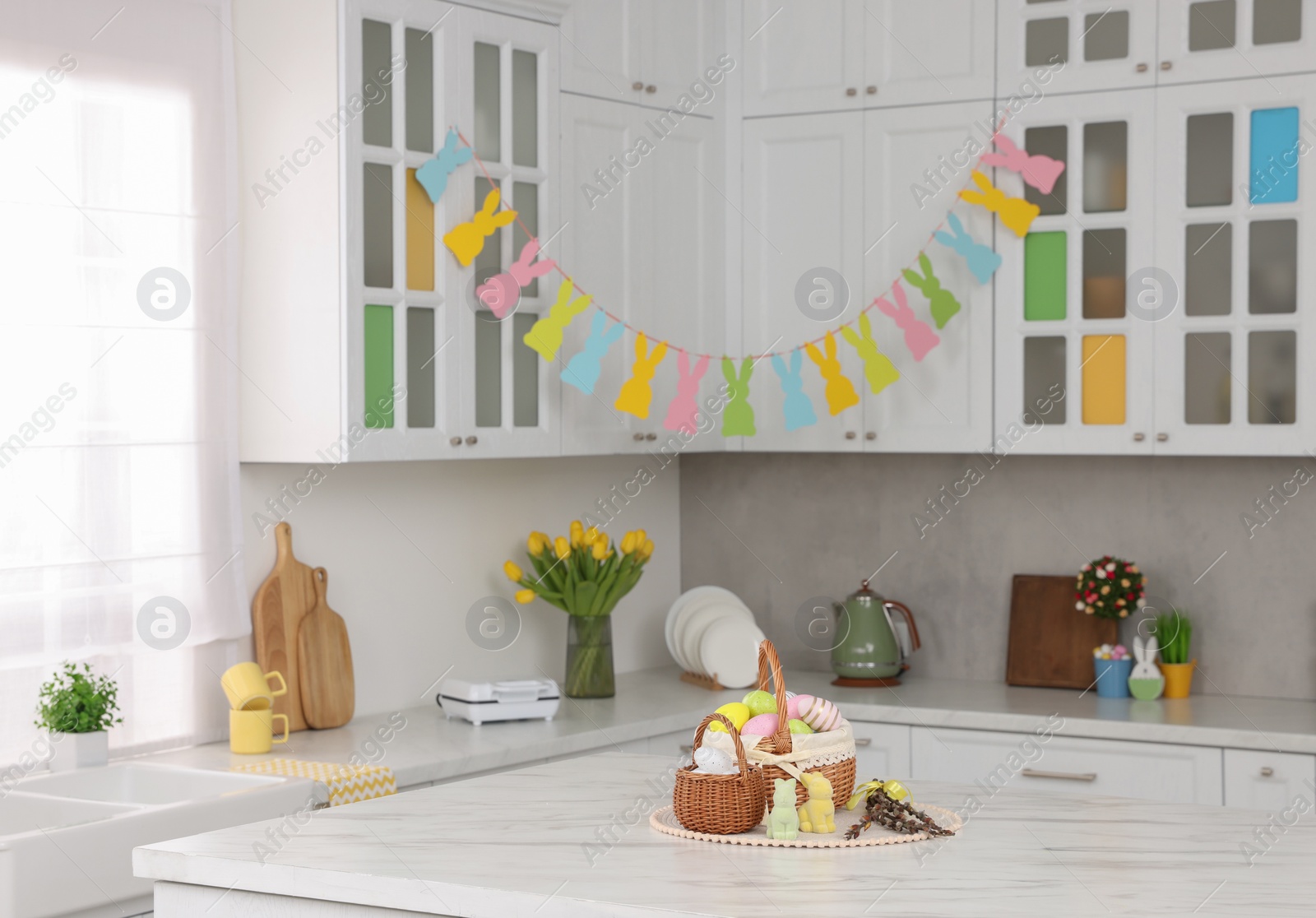 Photo of Wicker baskets with Easter eggs at white marble table and festive decor in kitchen