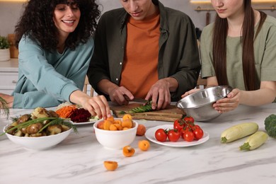 Photo of Friends cooking healthy vegetarian meal at white marble table in kitchen