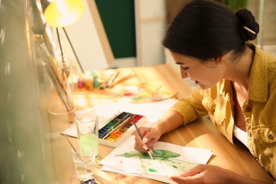 Photo of Young woman drawing leaf with watercolors at table indoors