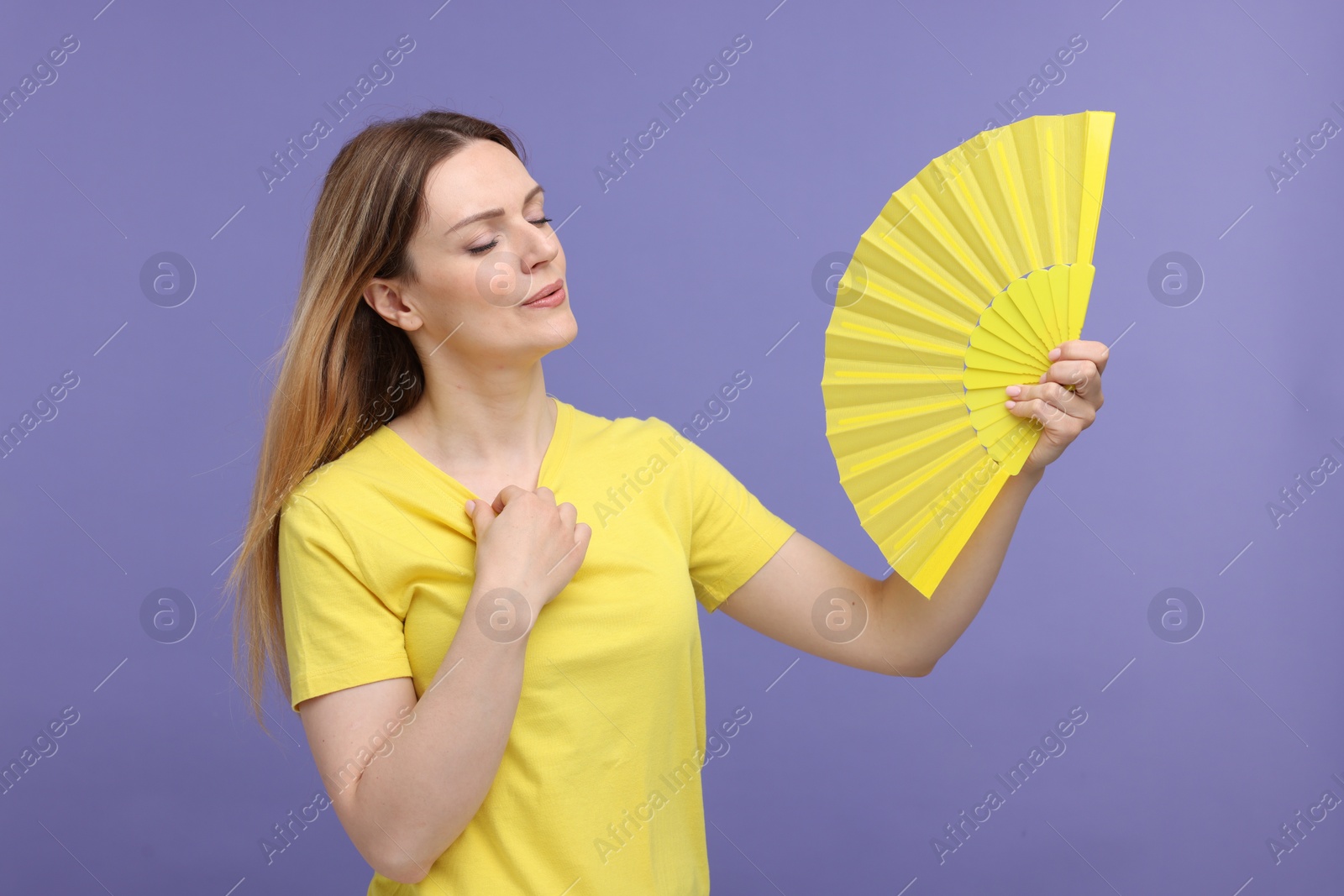 Photo of Beautiful woman waving yellow hand fan to cool herself on violet background