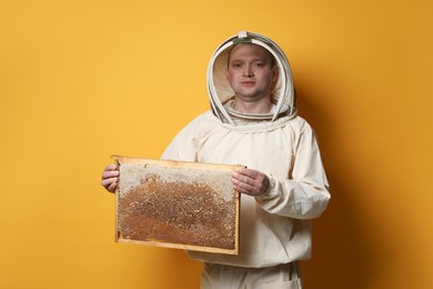 Photo of Beekeeper in uniform holding hive frame with honeycomb on yellow background