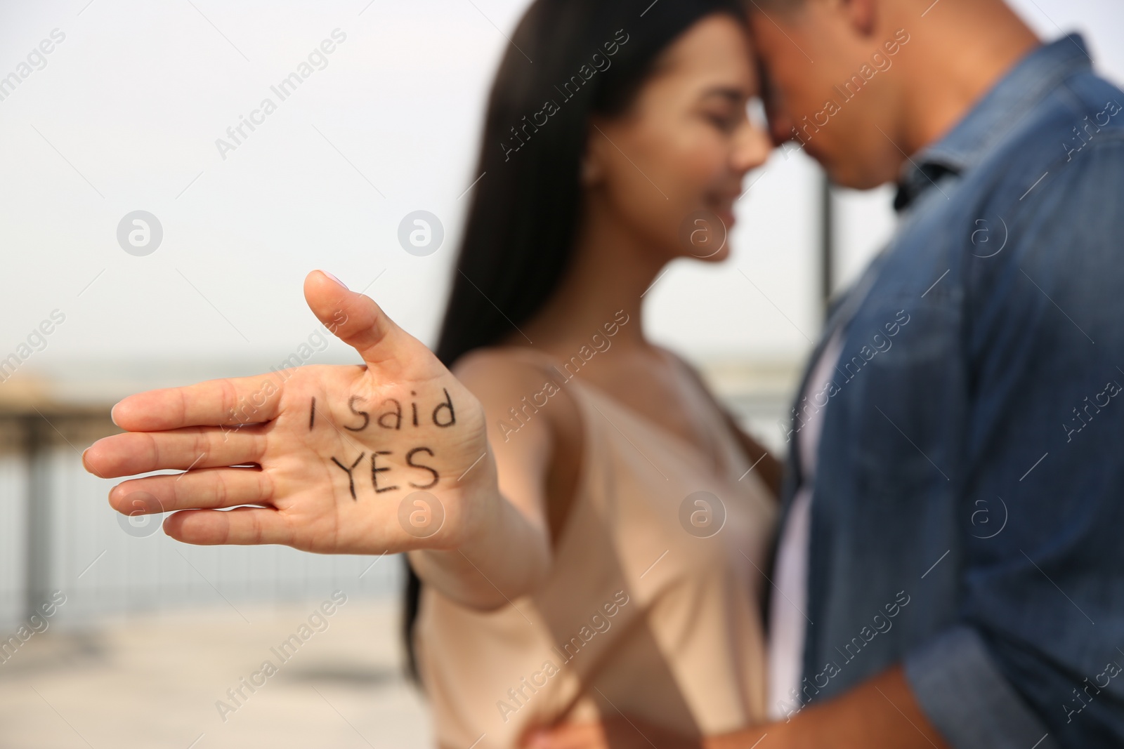 Photo of Woman with text I SAID YES written on palm and her boyfriend after engagement outdoors, focus on hand