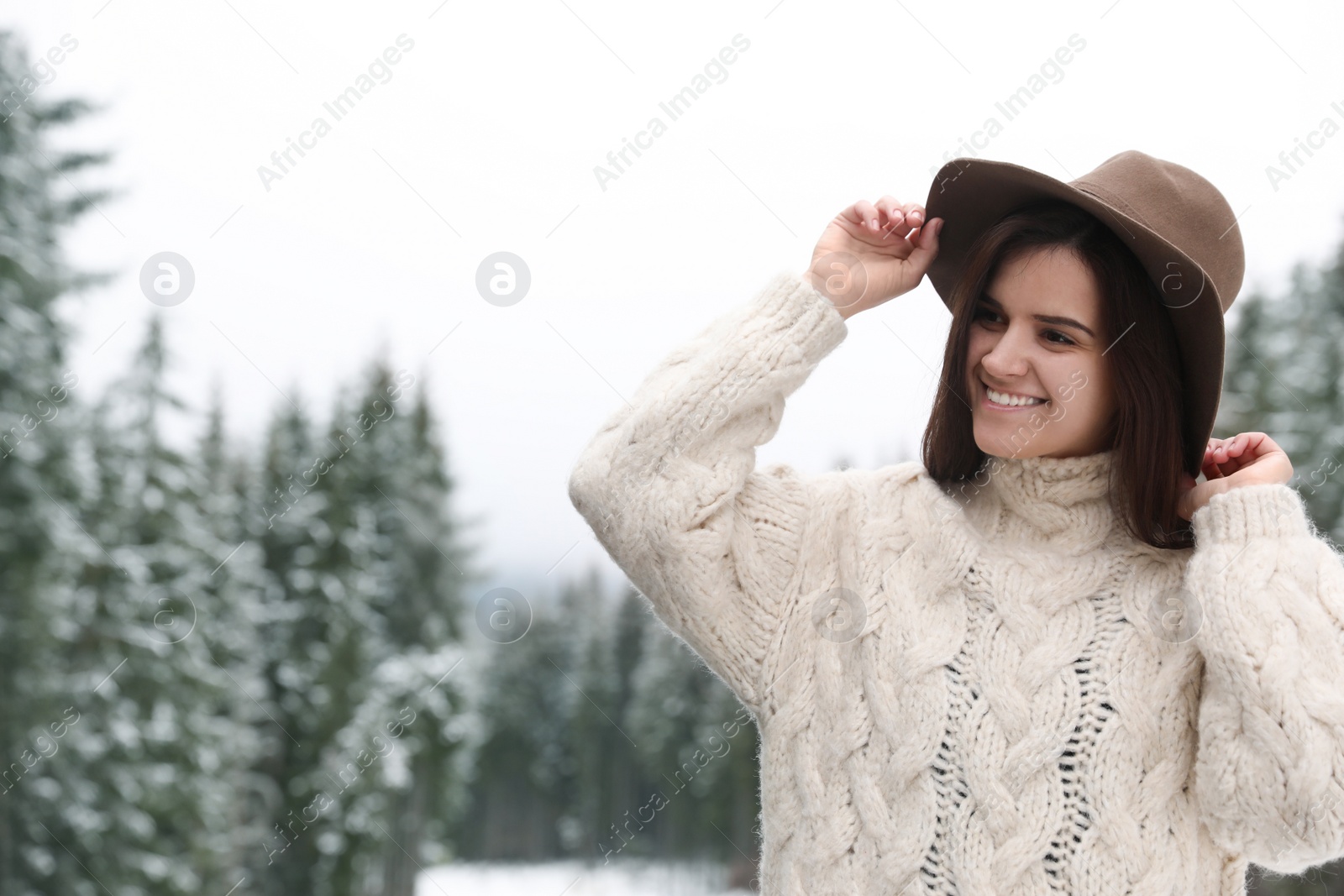 Photo of Young woman in warm sweater near snowy forest, space for text. Winter vacation