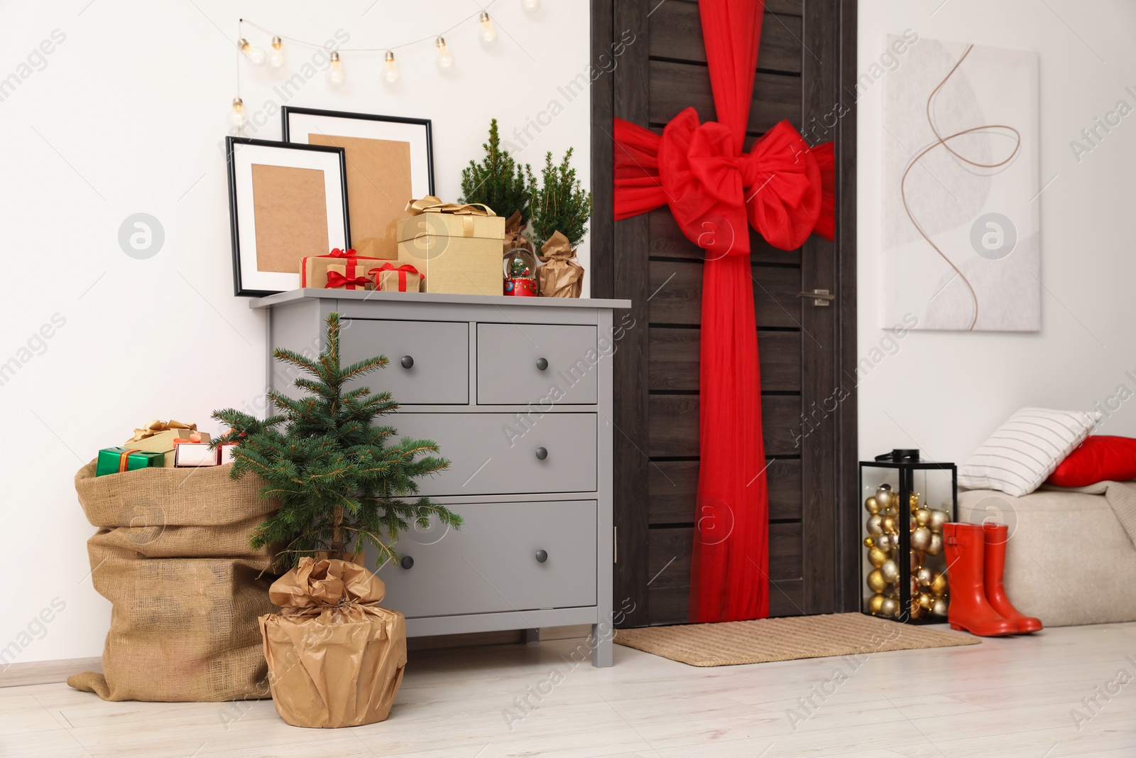 Photo of Beautiful fir tree and sack with Christmas gifts near chest of drawers and wooden door decorated with red bow in room