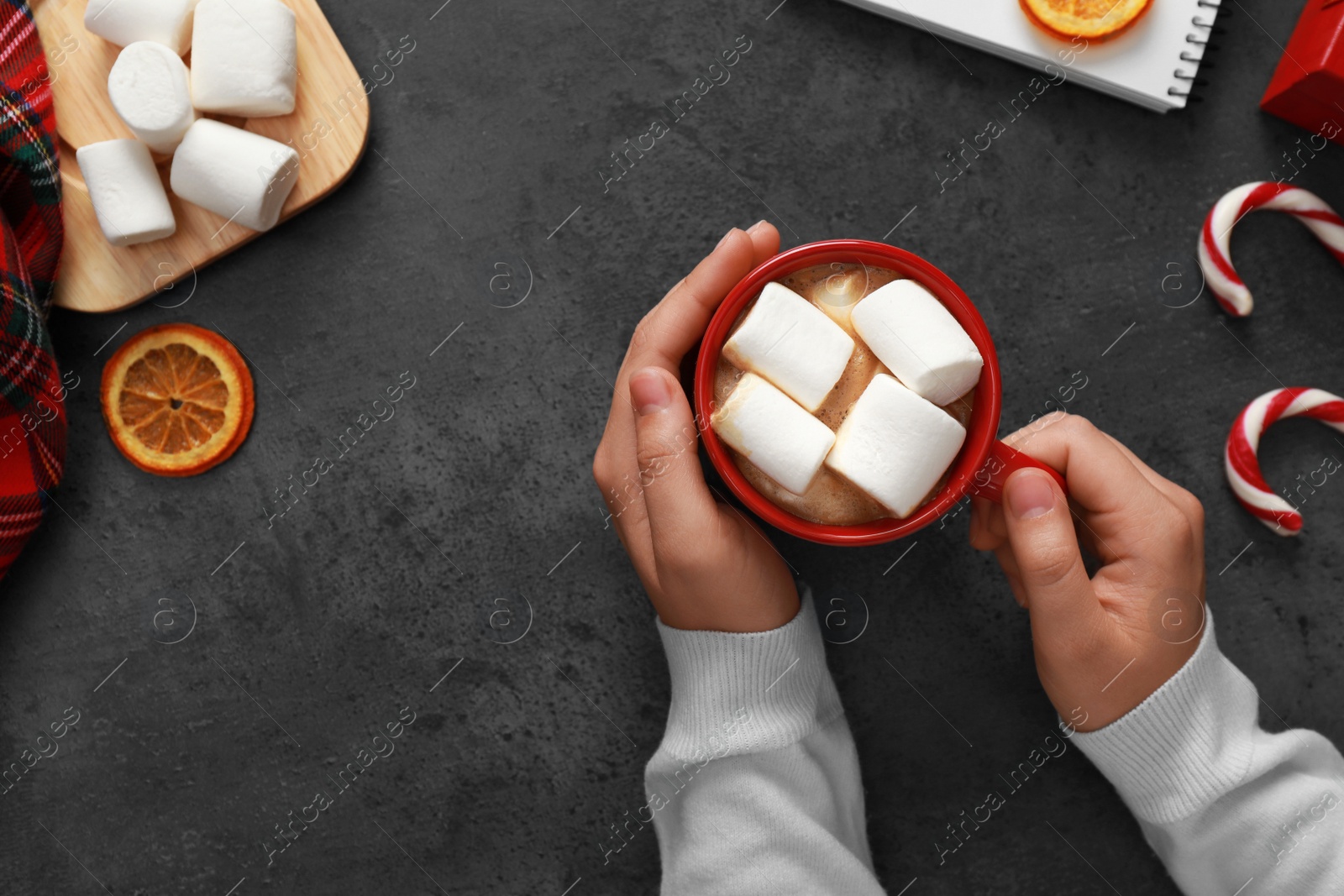 Photo of Woman holding cup of tasty cocoa with marshmallows at dark grey table, top view. Cozy winter