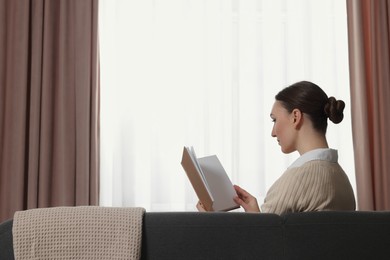 Woman reading book on sofa near window with stylish curtains at home. Space for text
