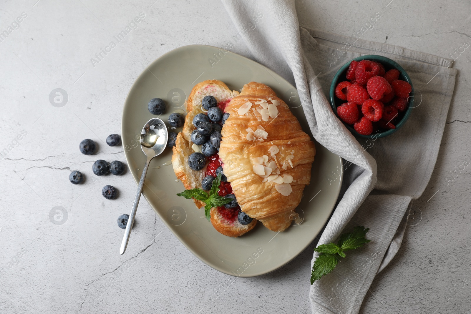 Photo of Delicious croissant with berries, almond flakes and spoon on light grey table, flat lay