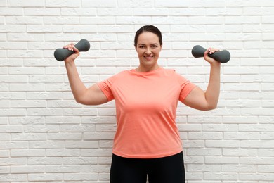 Photo of Happy overweight woman doing exercise with dumbbells near white brick wall
