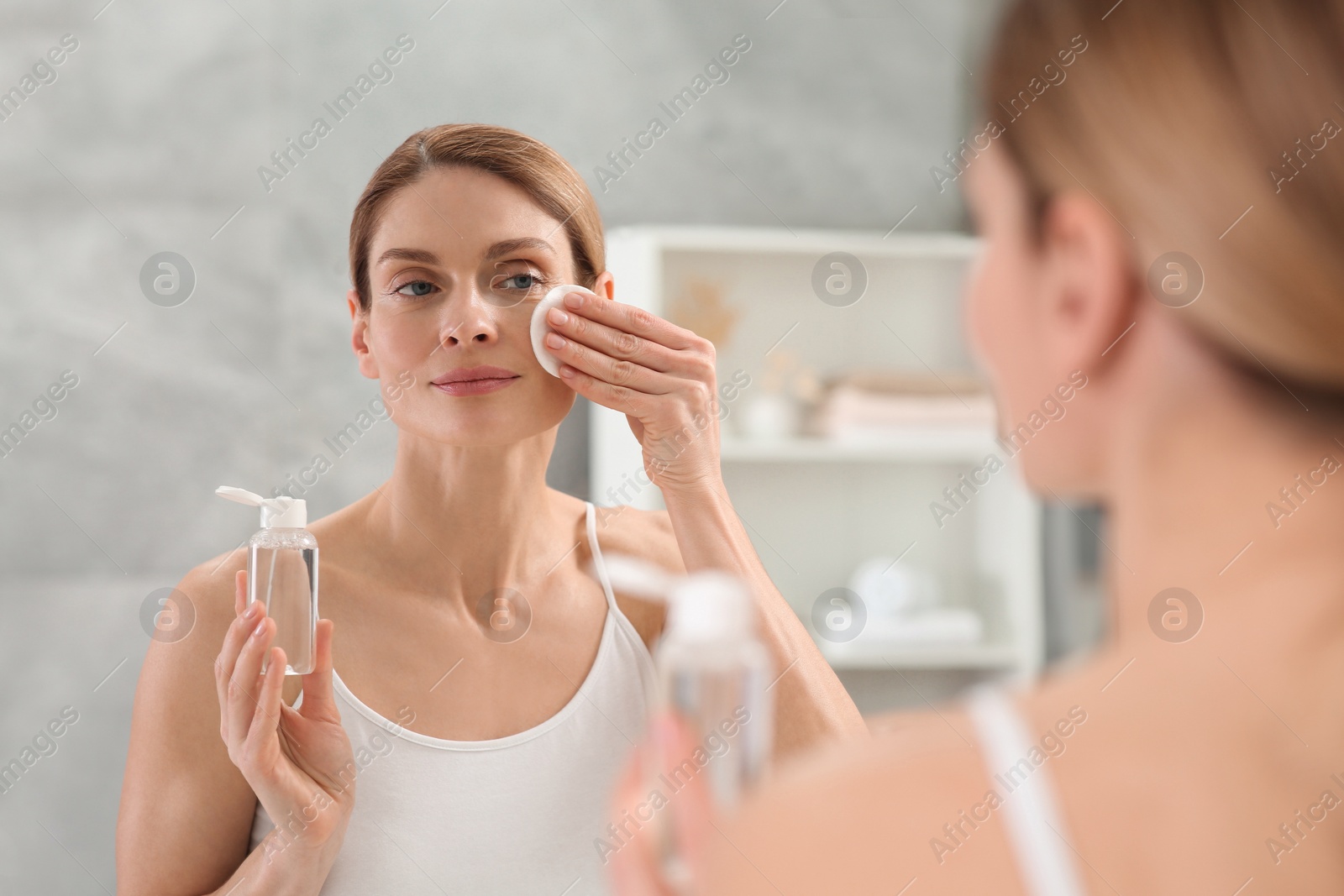 Photo of Beautiful woman removing makeup with cotton pad near mirror indoors