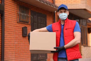 Courier in protective mask and gloves with box near house outdoors. Delivery service during coronavirus quarantine