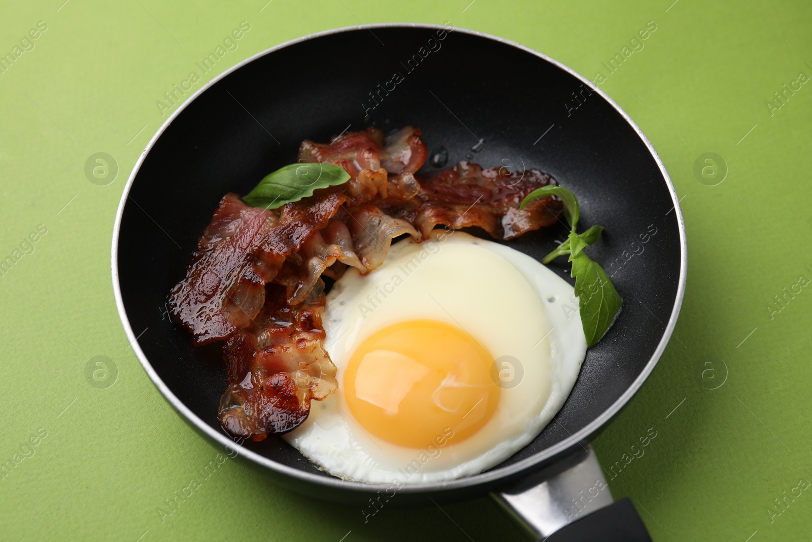 Photo of Fried egg, bacon and basil in frying pan on green background